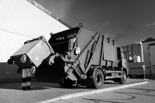 A large commercial waste collection truck is positioned on a paved street next to an industrial or commercial building with a high, corrugated metal wall. The truck has a hydraulic lifting mechanism and an enclosed container at the rear, which is filling with various types of rubbish. A worker wearing protective gloves is seen loading or inspecting the waste, which includes a large rectangular bin with a textured surface, possibly made of plastic or metal. The scene is set outdoors under a partly cloudy sky, illustrating the outdoor rubbish collection services provided by Garden Clearance Tottenham in the local area, possibly near Tottenham or the surrounding postcode district. The image captures the operational environment typical of outdoor rubbish removal activities, emphasizing the company’s expertise in handling commercial and industrial waste disposal efficiently.