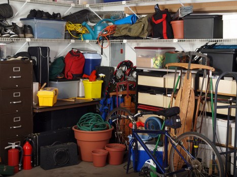 The image depicts a cluttered storage area or garage with various household items and outdoor equipment. On the left side, a tall black filing cabinet and a small yellow container are visible, along with a red backpack and a blue pet carrier placed on a workbench surface. Resting on the workbench are a yellow bucket and a coiled green garden hose in a terracotta pot. Behind, shelves are filled with assorted plastic containers, boxes, and camping equipment, including a tent and cables, extending across the wall. On the right side, a wooden hand truck leans against the shelving unit, with a small black toolbox nearby. A bicycle with a blue frame and black saddle is positioned in the foreground, partially blocking view of additional storage boxes and gardening tools. The environment appears to be a typical garage or shed space used for storing outdoor gear, tools, and miscellaneous household rubbish, reflecting typical clutter that may be addressed during rubbish removal services, such as those offered by Garden Clearance Tottenham in the Tottenham area of London.