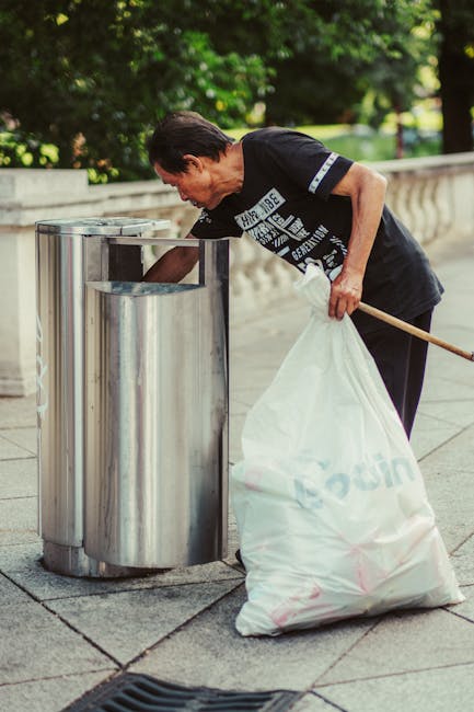 Close-up view of a person dressed in bright orange work overalls and wearing a white glove, holding a blue plastic rubbish bag filled with waste material. The bag is tightly gathered at the top, with 