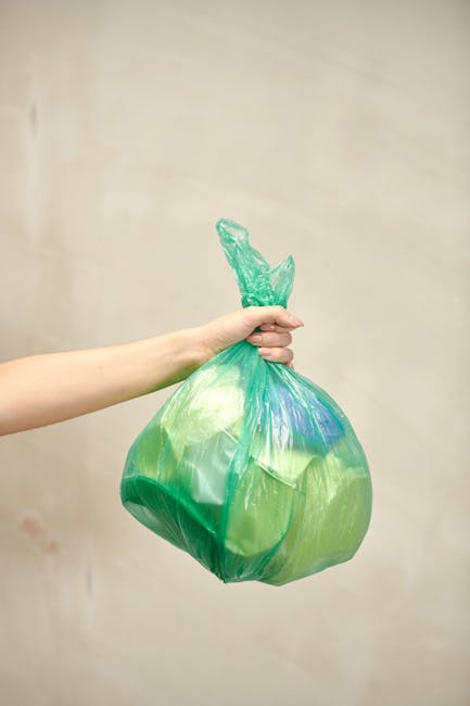 A person's arm is extended into the frame from the left, holding a green plastic rubbish bag tightly by the tied top. The bag appears to be filled with waste material, creating a rounded shape with vi